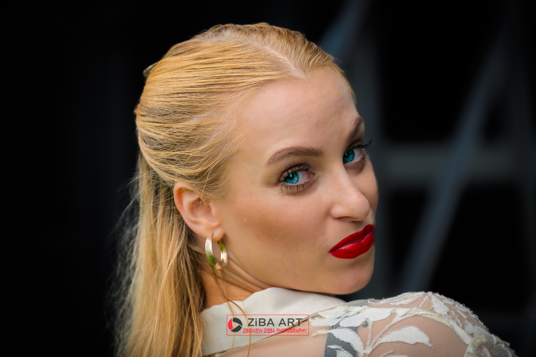 Portret kobiety z blond włosami, intensywnym makijażem oczu i czerwonymi ustami, spoglądającej przez ramię, z widocznym logo fotografa w rogu.