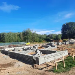 Wznoszenie fundamentów z bloczków betonowych na piaszczystym podłożu, widoczne wystające pręty zbrojeniowe i stos bloczków w tle, błękitne niebo.