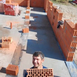 THE BEST ESTATES SPÓŁKA Z OGRANICZONĄ ODPOWIEDZIALNOŚCIĄ - Uśmiechnięty murarz prezentuje pustak ceramiczny Wienerberger Porotherm na budowie domu, widoczne wznoszone ściany z pustaków i zapasy materiału.