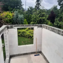 Wnętrze wznoszonej konstrukcji z białych bloczków, widok na zielony ogród przez otwór okienny, na podłodze leży miotła.