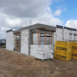 Surowy stan budowy domu z białych bloczków Ytong, widoczne zbrojenia i drewniane elementy konstrukcyjne, paleta bloczków i zapakowane paczki Ytong na placu budowy.