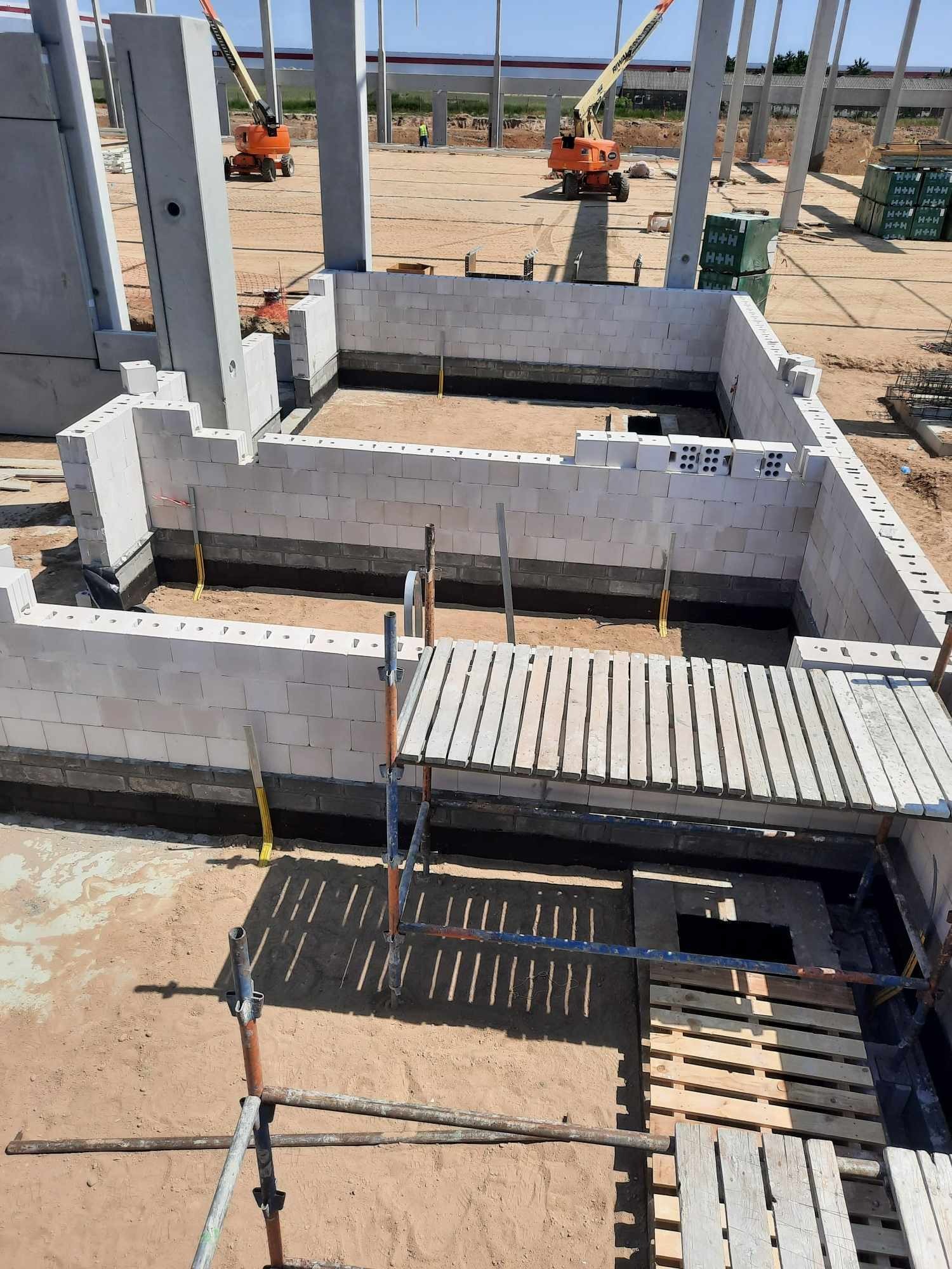 Wznoszenie ścian z bloczków betonowych na fundamencie, widoczne rusztowanie i słupy konstrukcyjne, w tle podnośniki budowlane.