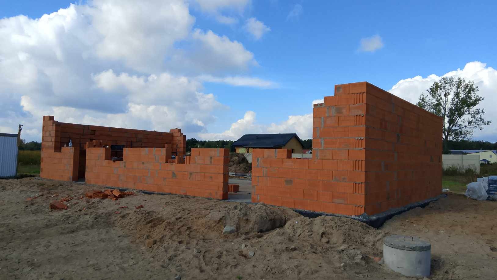 Wznoszone ściany z czerwonego bloczka na fundamencie nowo budowanego domu jednorodzinnego, widoczne fragmenty zbrojenia i sąsiedni budynek w tle.