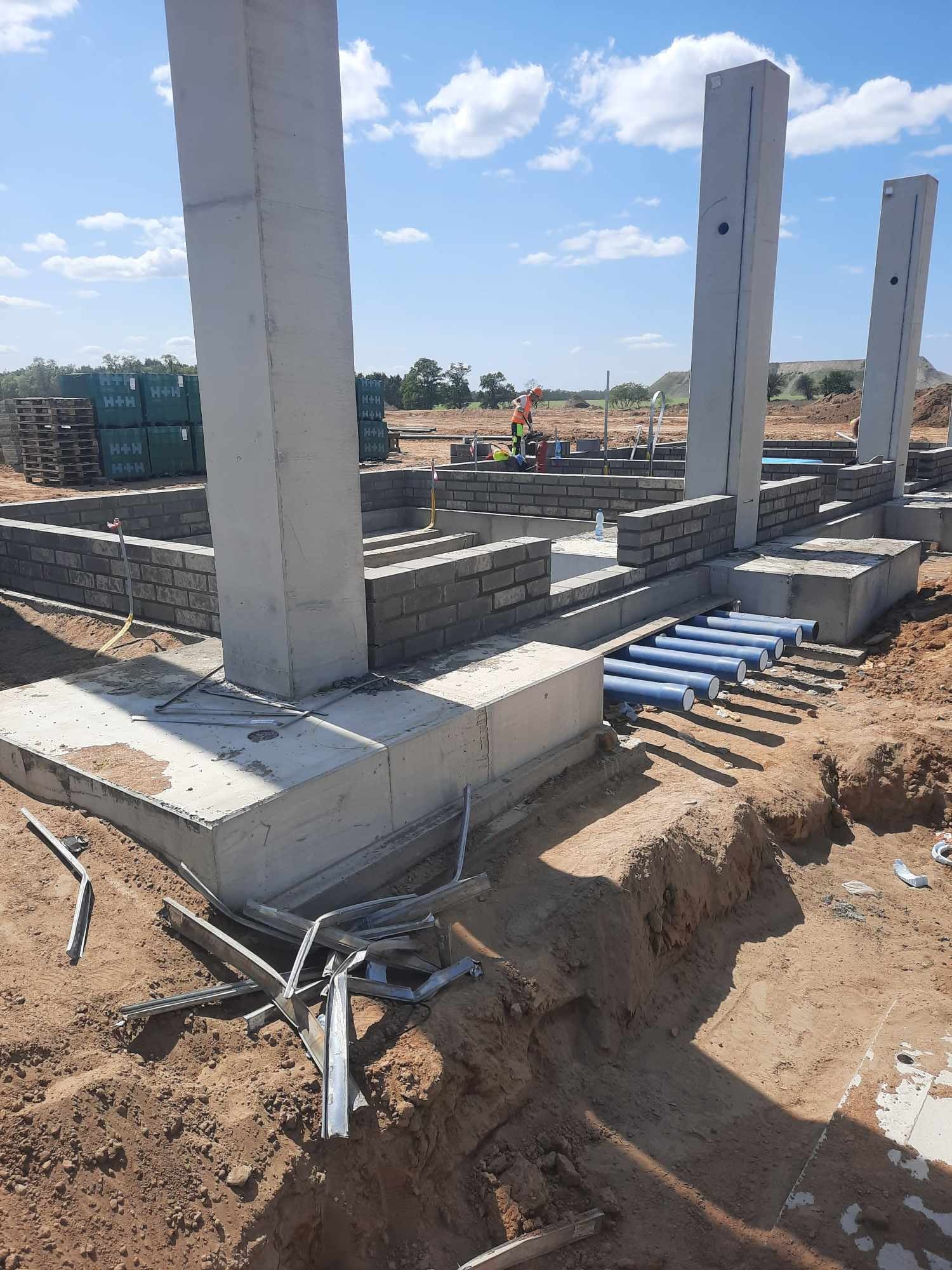 Wznoszenie fundamentów i ścian z bloczków betonowych, widoczne pionowe słupy konstrukcyjne oraz niebieskie rury instalacyjne w wykopie, w tle palety z bloczkami H+H.