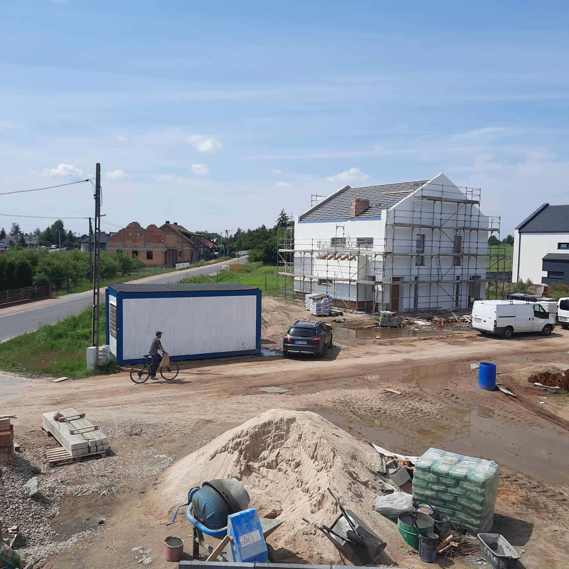 Wznoszony dom murowany ze szkieletem rusztowania, widoczny na tle błękitnego nieba, z kupą piasku, betoniarką i paletami z cegłami na pierwszym planie, a obok kontener budowlany i rowerzysta.