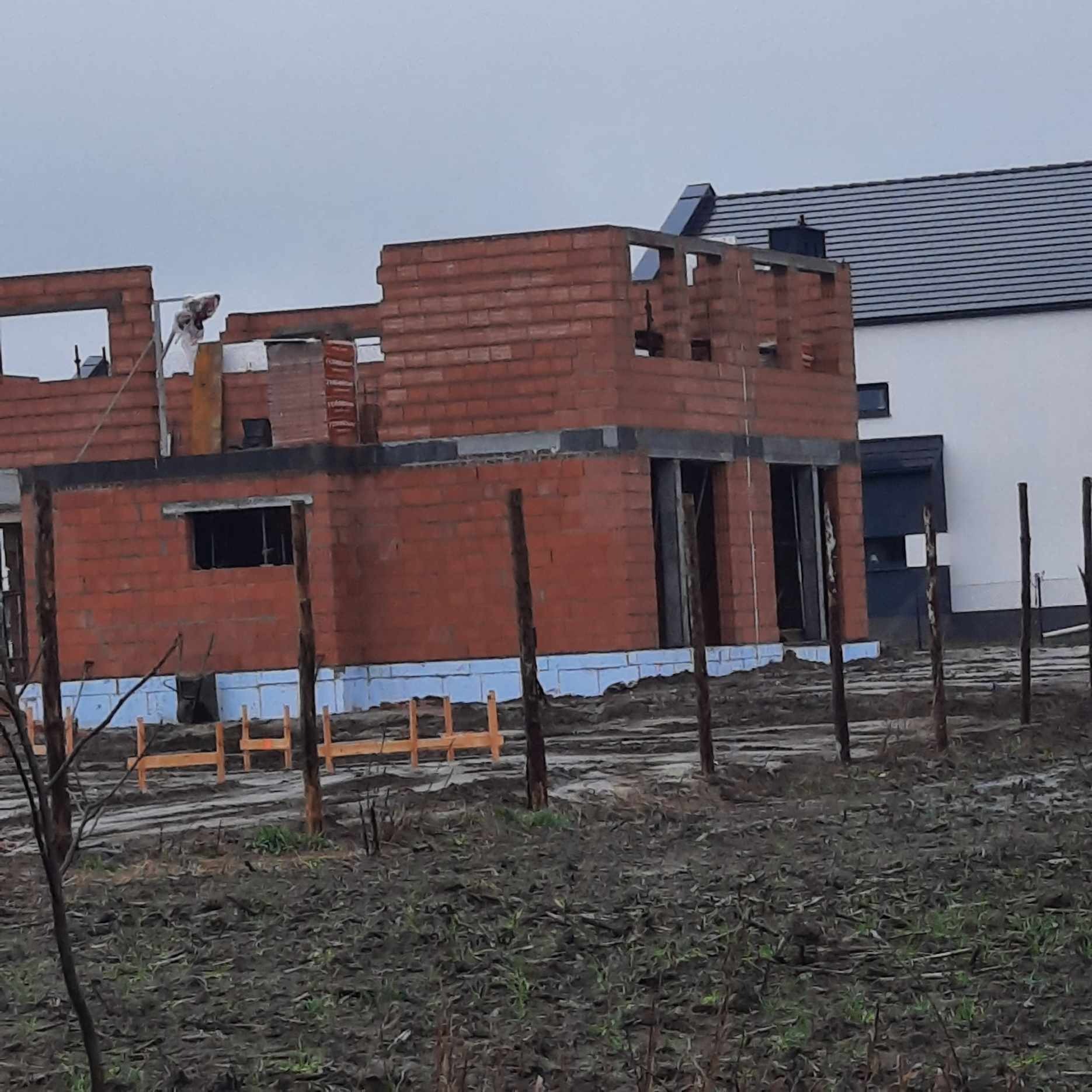 Surowy stan budowy domu z czerwonej cegły, widoczne drewniane szalunki i fundamenty, w tle biały budynek z ciemnym dachem, pochmurne niebo.