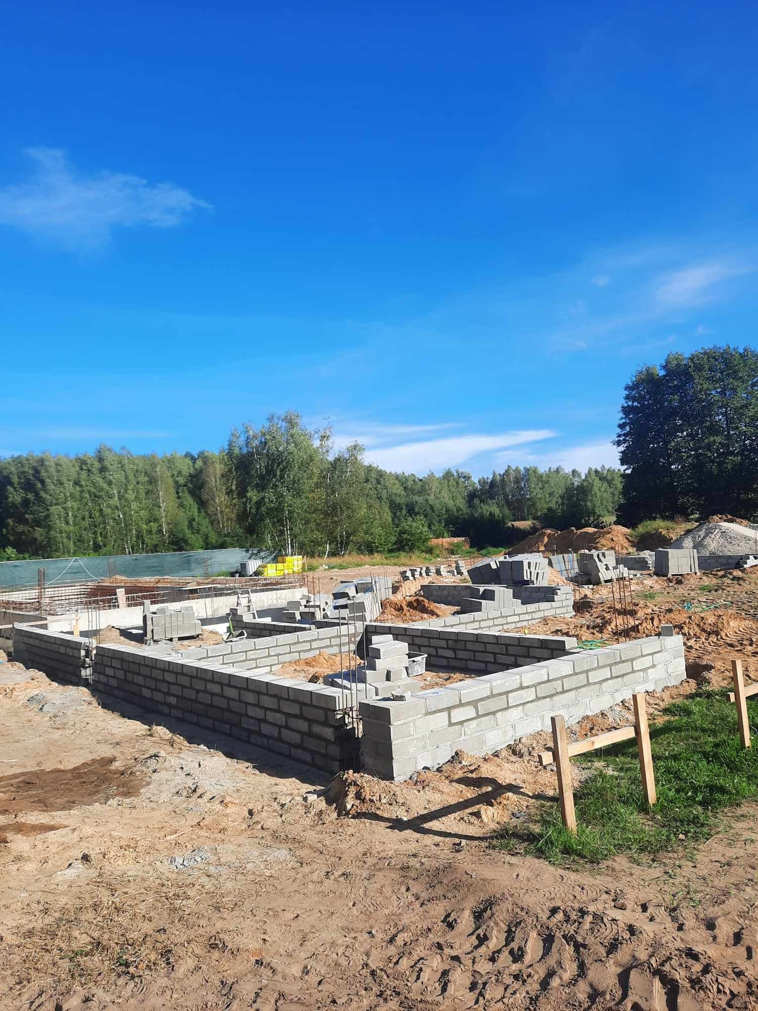 Wznoszenie fundamentów z bloczków betonowych na piaszczystym podłożu, widoczne wystające pręty zbrojeniowe i stos bloczków w tle, błękitne niebo.