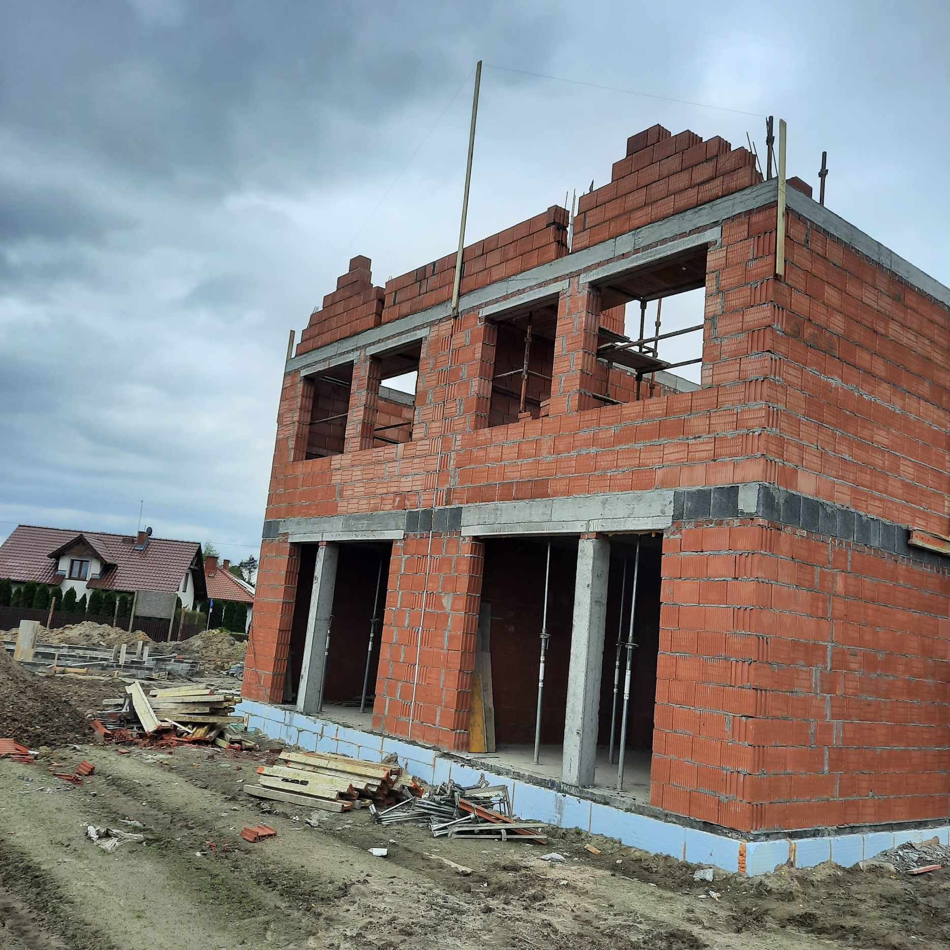 Surowy stan dwukondygnacyjnego domu z czerwonej cegły, widoczne otwory okienne i drzwiowe, szalunki stropowe, drewniane podpory, materiały budowlane na ziemi, w tle dom jednorodzinny.