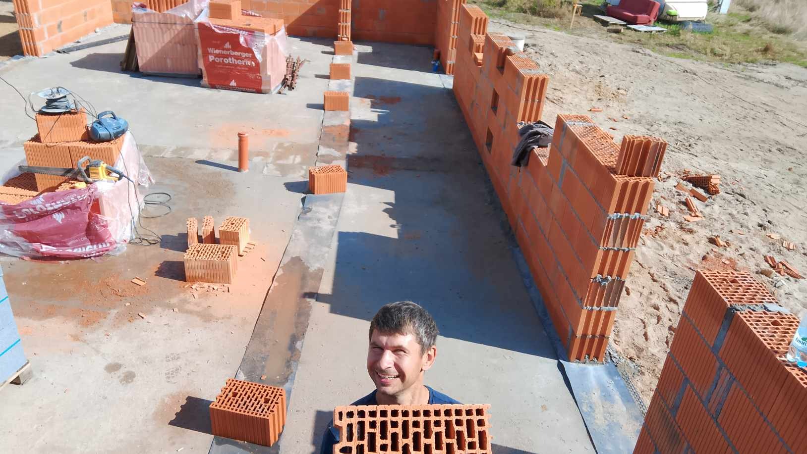 Uśmiechnięty murarz prezentuje pustak ceramiczny Wienerberger Porotherm na budowie domu, widoczne wznoszone ściany z pustaków i zapasy materiału.