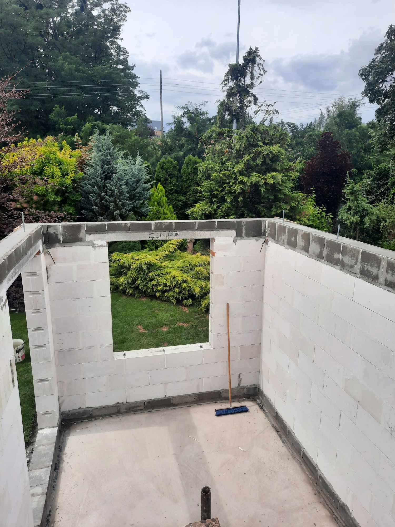 Wnętrze wznoszonej konstrukcji z białych bloczków, widok na zielony ogród przez otwór okienny, na podłodze leży miotła.