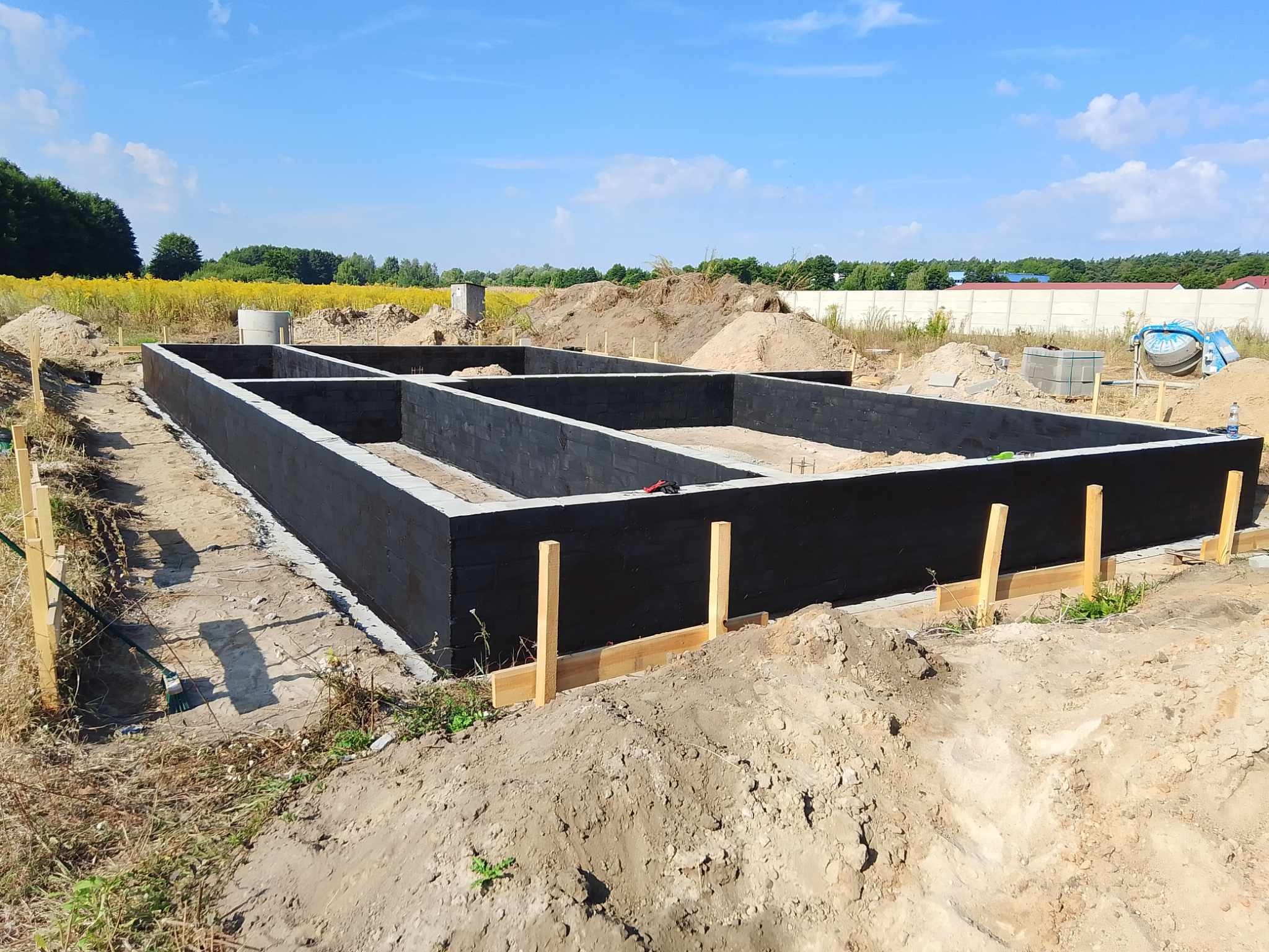 Zalane fundamenty pod dom z bloczków betonowych, zabezpieczone izolacją pionową, widoczne szalunki, w tle pole z kwitnącymi żółtymi kwiatami.