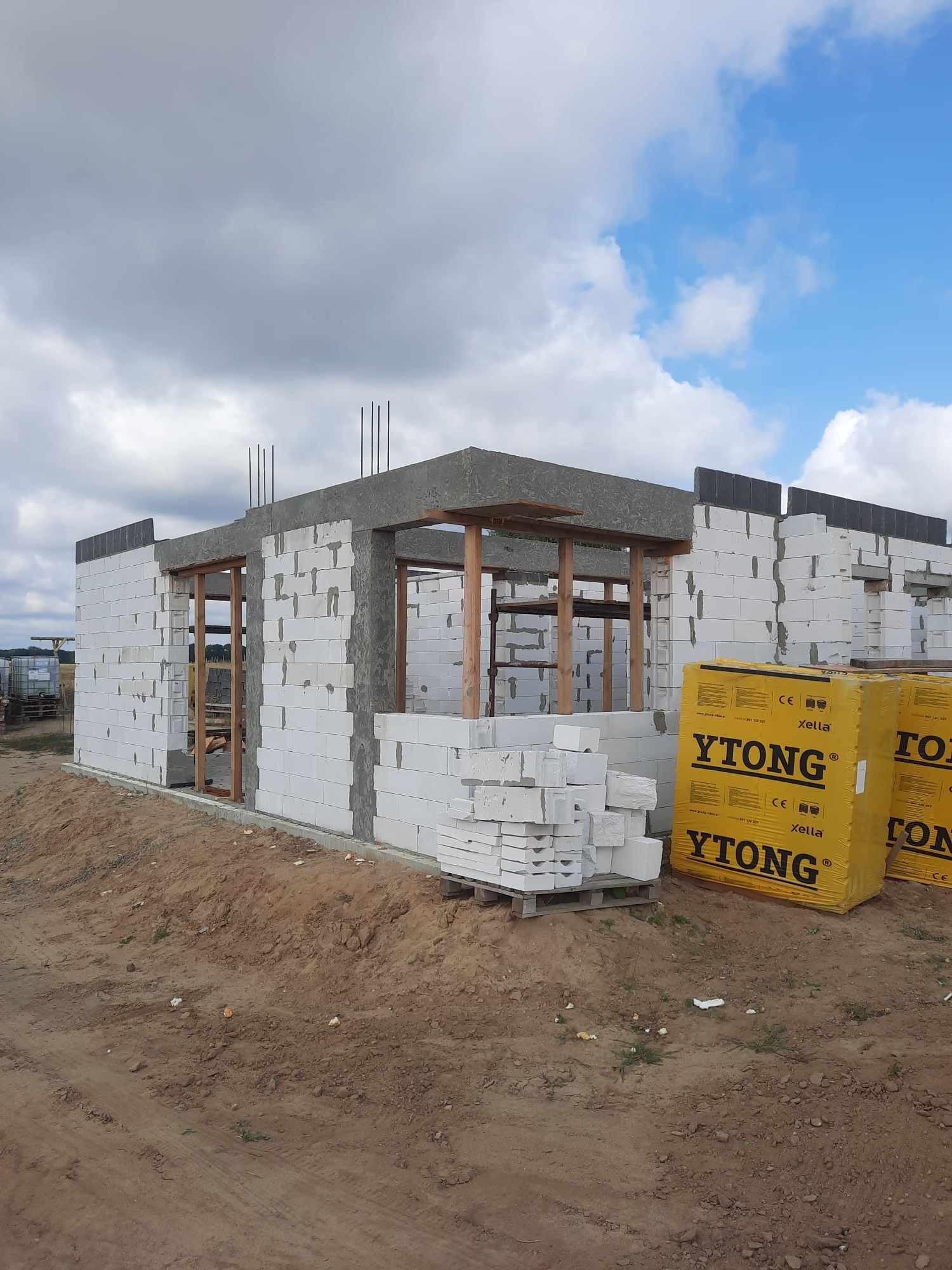 Surowy stan budowy domu z białych bloczków Ytong, widoczne zbrojenia i drewniane elementy konstrukcyjne, paleta bloczków i zapakowane paczki Ytong na placu budowy.