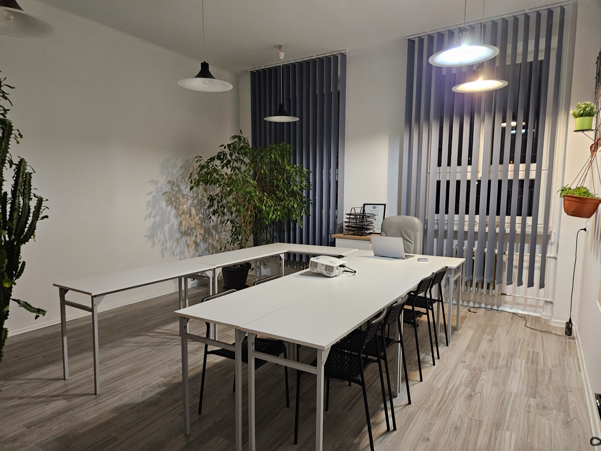Jasne biuro z białymi stołami ułożonymi w kształt litery U, czarnymi krzesłami, projektorem i laptopem na stole, roletami pionowymi w oknach i roślinami doniczkowymi.