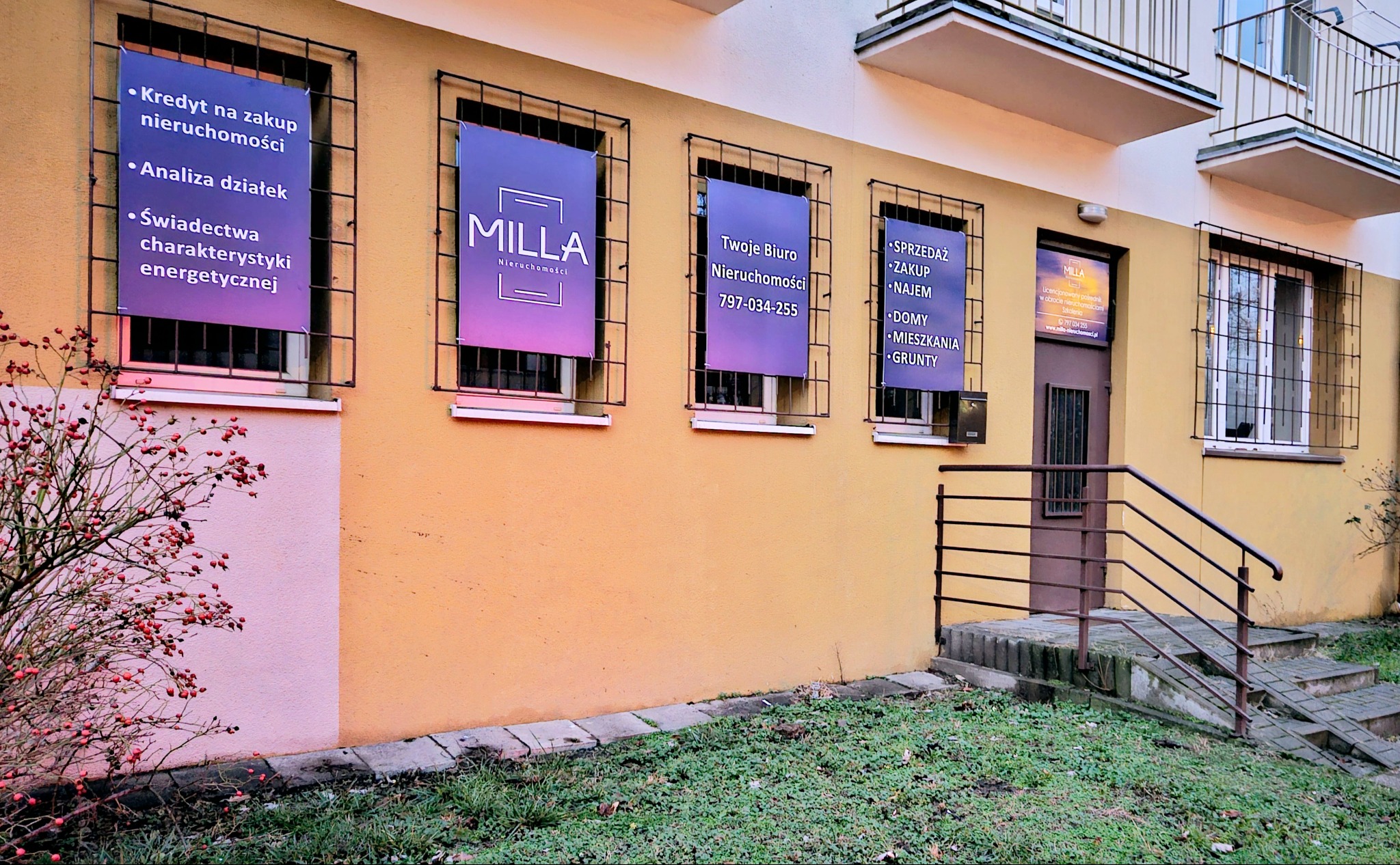 Fasada biura nieruchomości Milla w Łodzi z plakatami w oknach, oferującymi kredyty na zakup nieruchomości, analizę działek, świadectwa charakterystyki energetycznej, sprzedaż, zakup i wynajem domów...