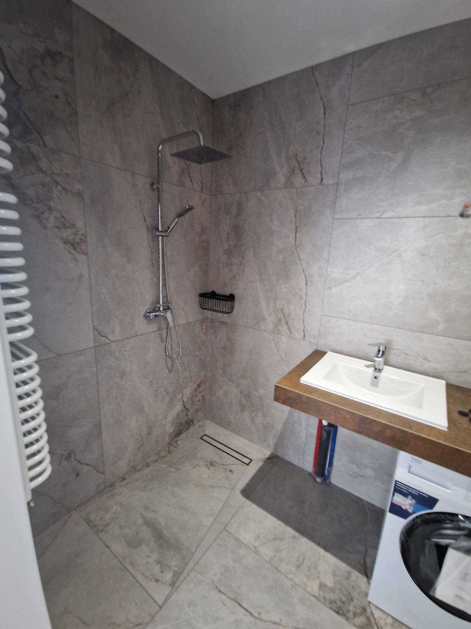 Wykończona łazienka z kabiną prysznicową typu walk-in, płytkami imitującymi marmur na ścianach i podłodze, umywalką nablatową na drewnianym blacie oraz pralką pod umywalką, widoczny grzejnik...