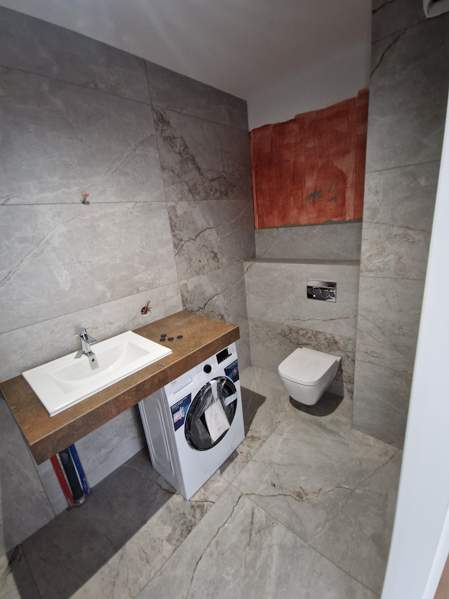 Łazienka w trakcie remontu: szare płytki imitujące marmur na ścianach i podłodze, umywalka nablatowa na blacie zintegrowanym z pralką, wisząca toaleta, ściana częściowo przygotowana pod malowanie.