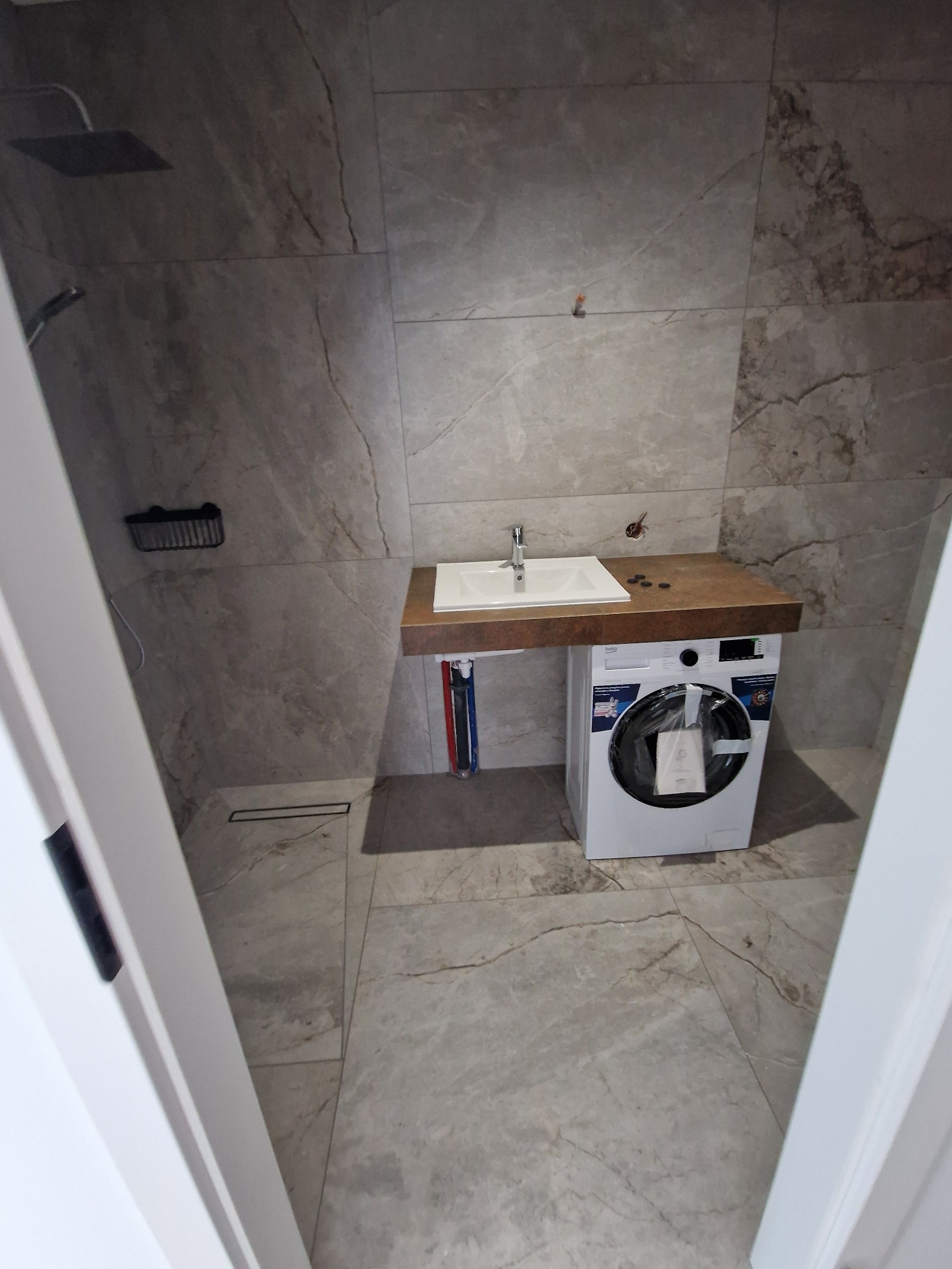 Wykończona łazienka z umywalką nablatową na drewnianym blacie, pod którym znajduje się pralka. Na ścianach i podłodze szare płytki imitujące marmur. Widoczny odpływ liniowy pod prysznicem.