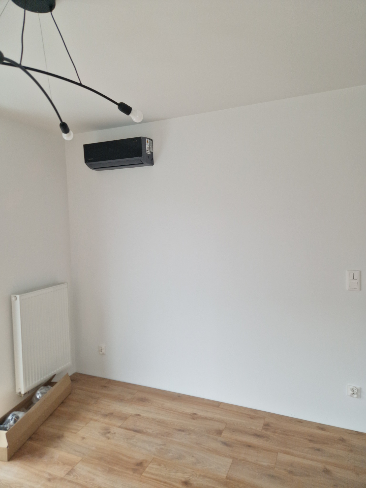 Wnętrze pokoju po remoncie: jasne panele podłogowe, białe ściany z zamontowanym czarnym klimatyzatorem i grzejnikiem, nowoczesna lampa sufitowa.