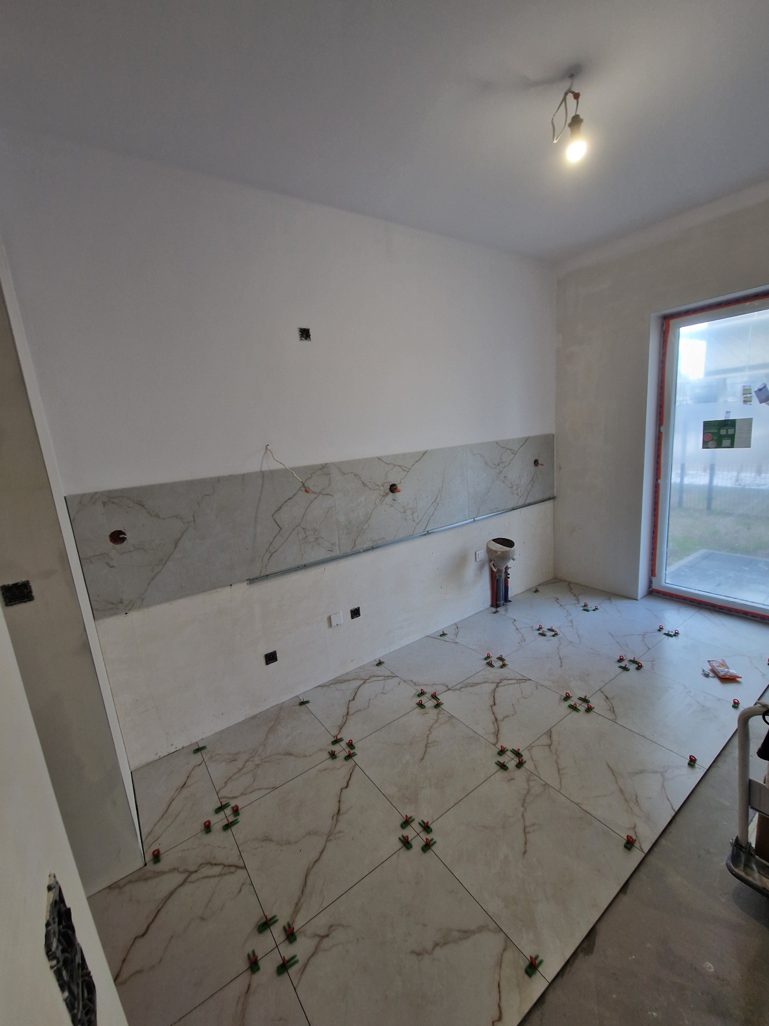 Układanie marmurowych płytek podłogowych w jasnym wnętrzu, z użyciem zielonych krzyżyków dystansowych, częściowo wykończona ściana z tym samym wzorem płytek.