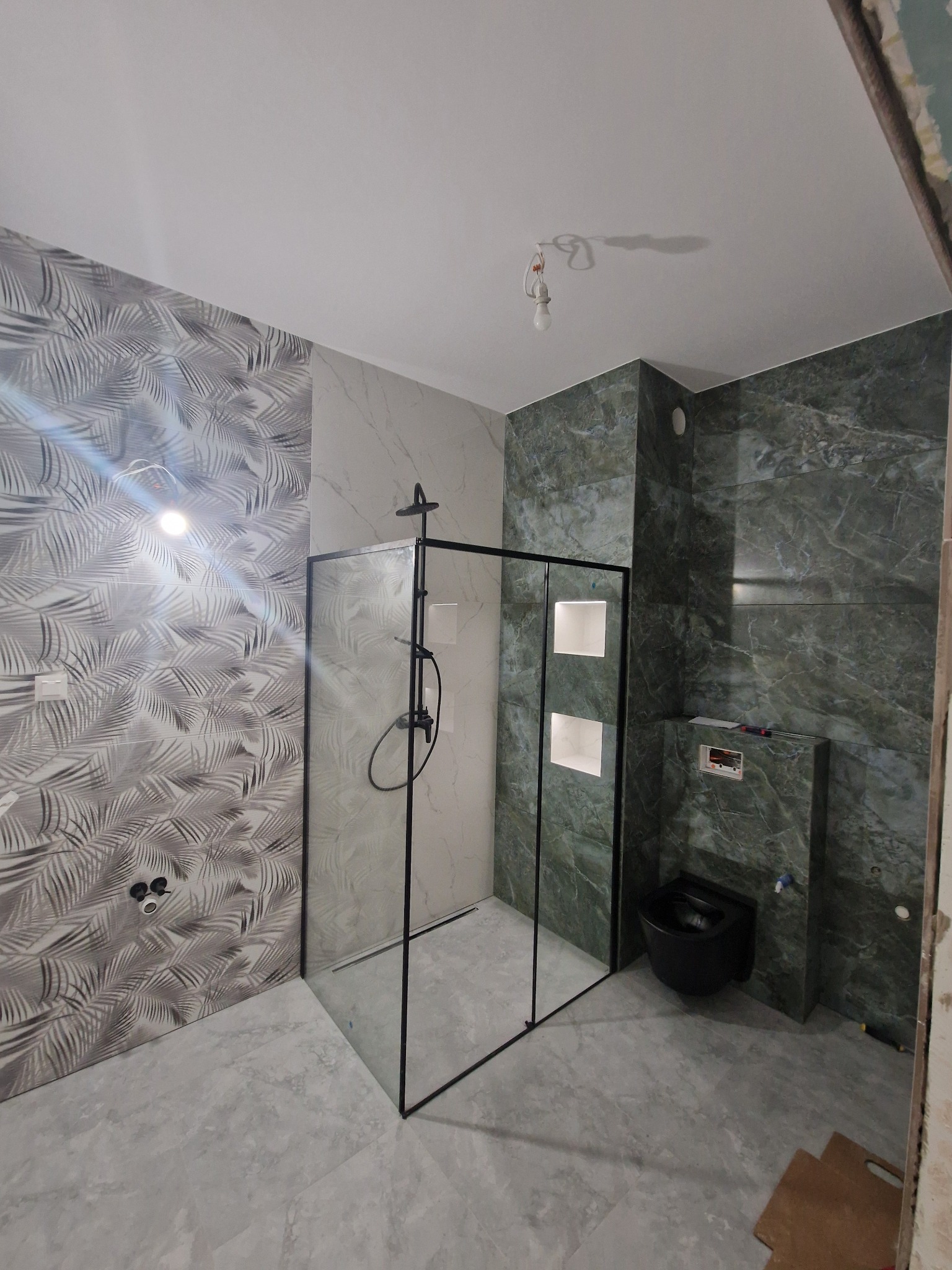 Wykończona łazienka z kabiną prysznicową typu walk-in z czarnymi profilami, wiszącą toaletą w kolorze czarnym oraz zróżnicowanymi wzorami płytek na ścianach: motyw liści palmowych, imitacja marmuru...
