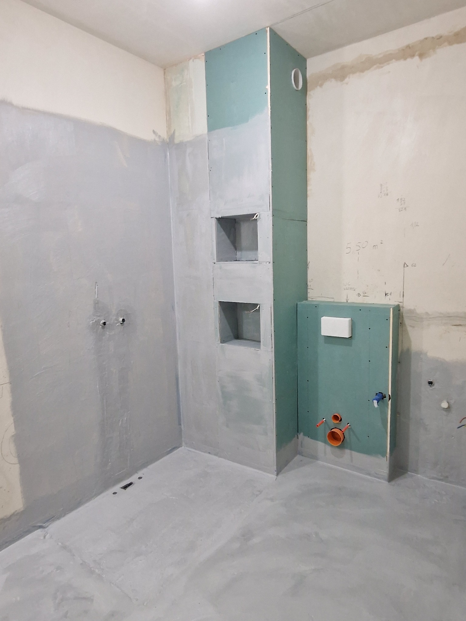 Surowe wnętrze łazienki w trakcie prac wykończeniowych: ściany częściowo pokryte szarą gładzią, zabudowa z płyt kartonowo-gipsowych z wnękami i instalacją sanitarną, widoczne rury i otwory...