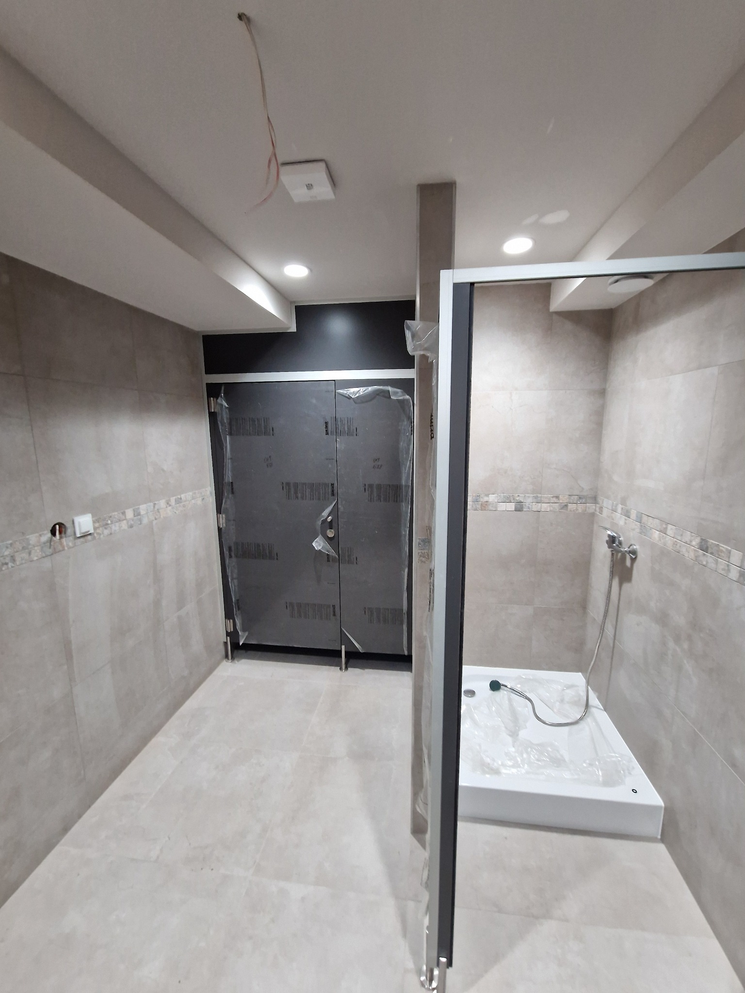 Wykończona łazienka w jasnych odcieniach szarości z kabiną prysznicową i drzwiami zabezpieczonymi folią ochronną, widoczne płytki ceramiczne na ścianach i podłodze z dekoracyjnym pasem płytek...