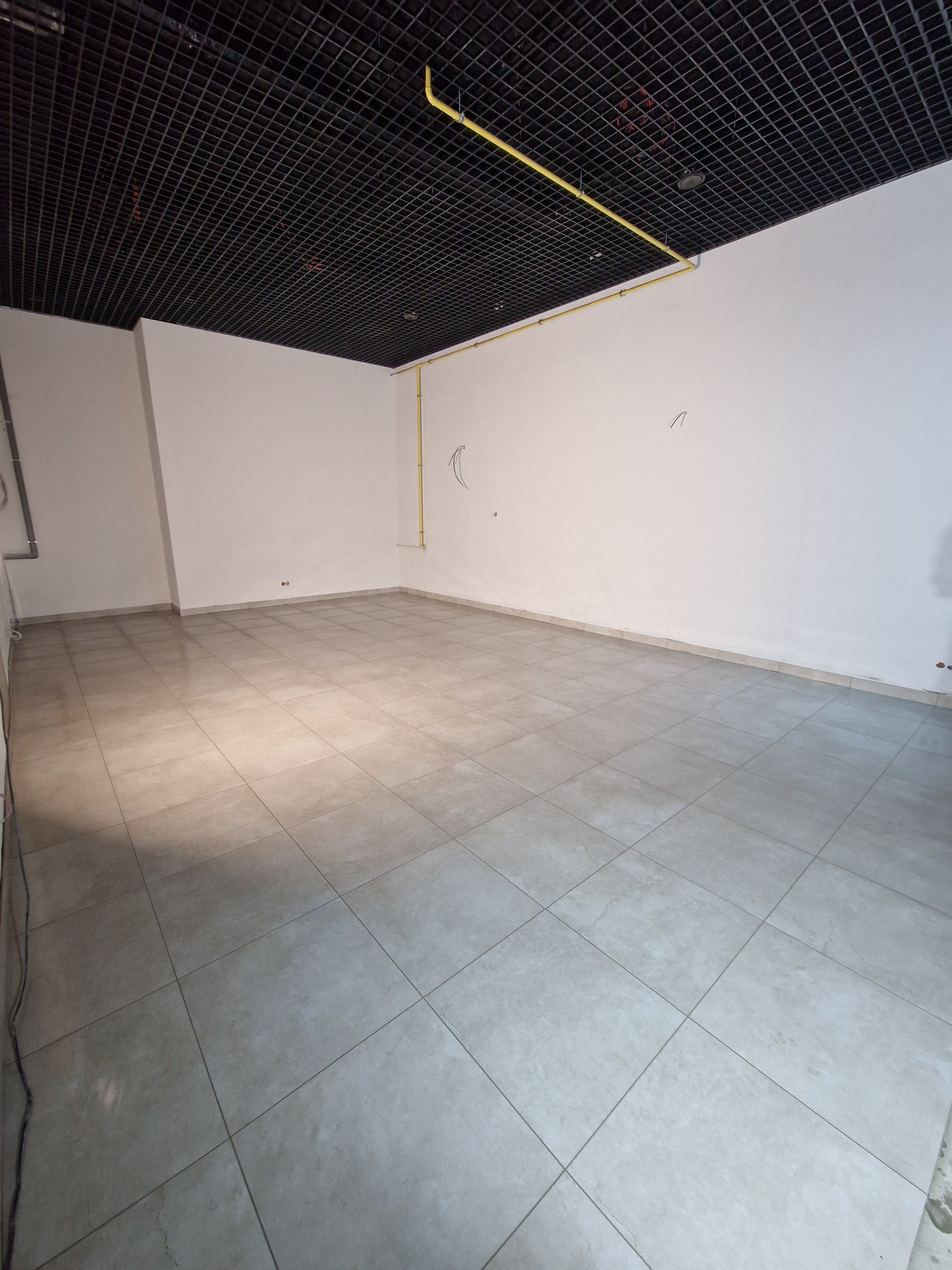 Wnętrze pustego pomieszczenia w Stryszawie z szarą, kwadratową podłogą z płytek, białymi ścianami i czarnym, kratowym sufitem podwieszanym z widocznymi żółtymi rurami instalacji.