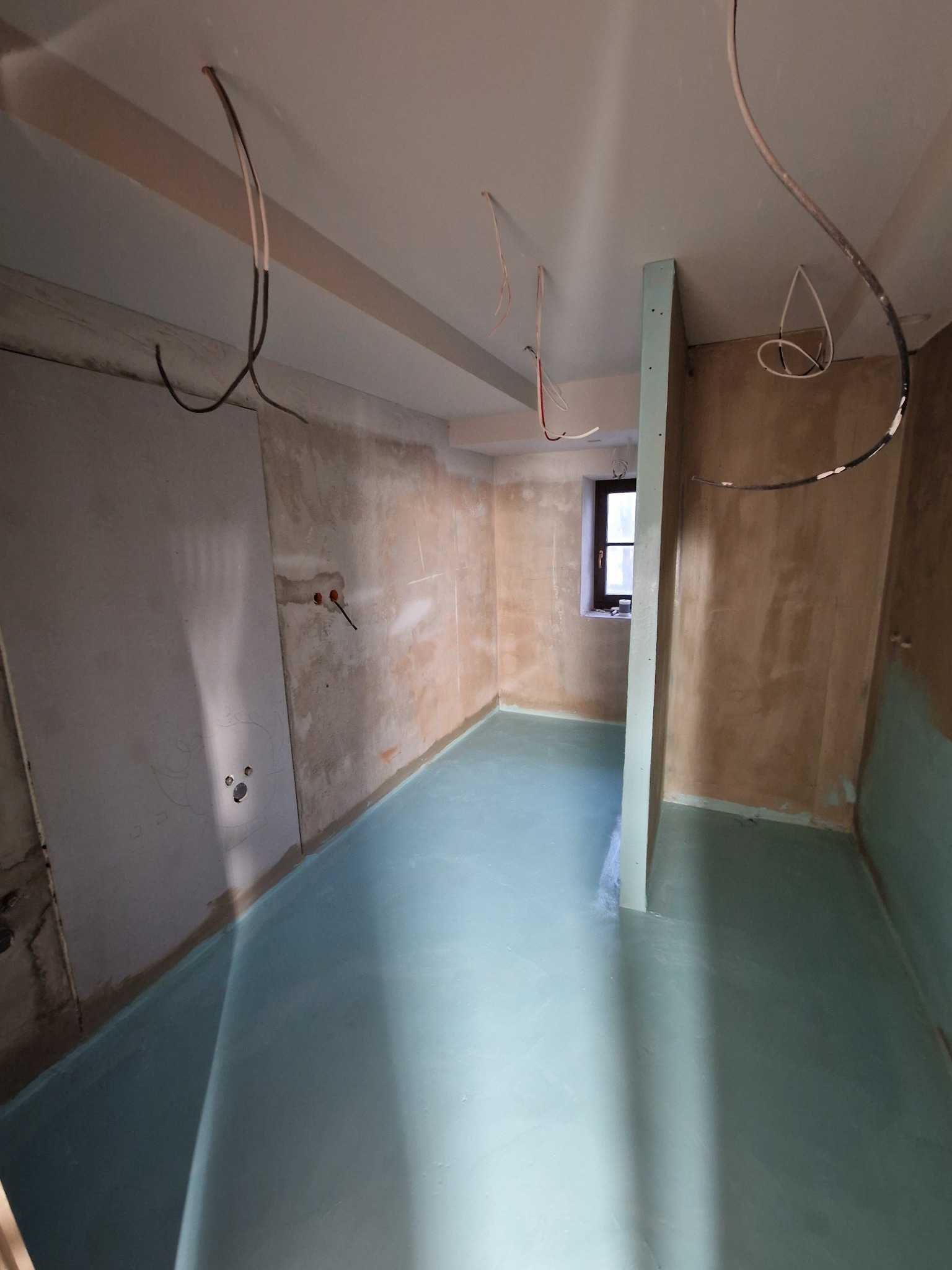 Przygotowane do prac wykończeniowych pomieszczenie z wylaną, jasnoniebieską posadzką samopoziomującą; ściany częściowo otynkowane, widoczne przewody elektryczne i fragment okna.