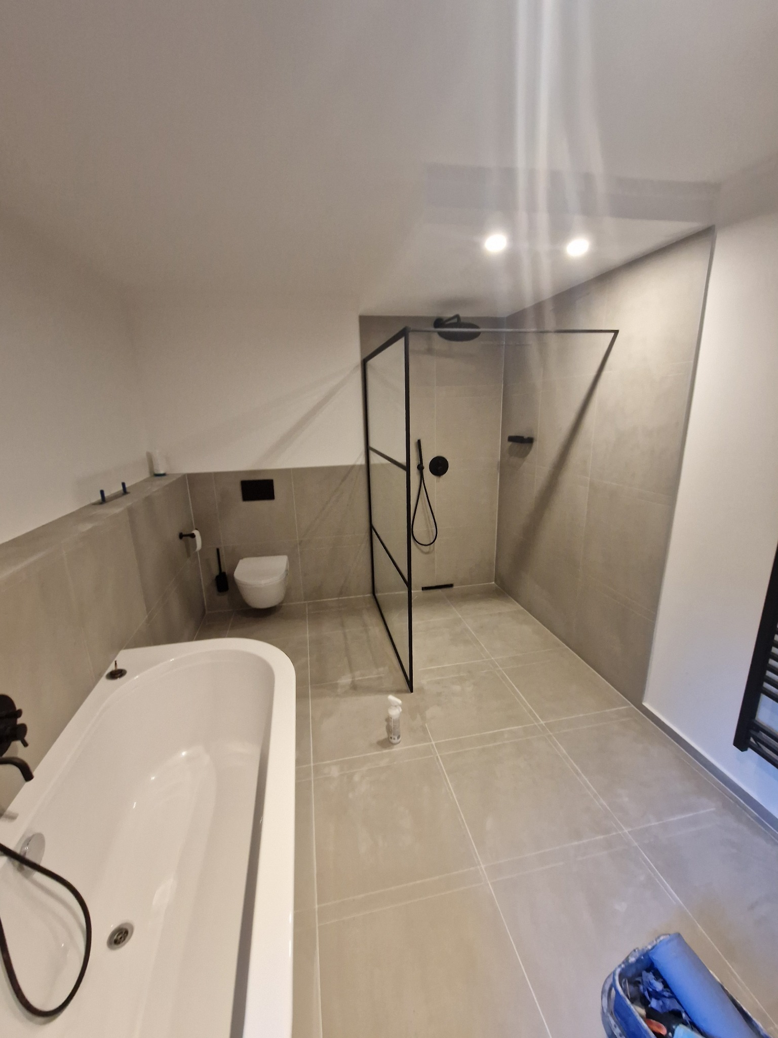 Wykończona łazienka z beżowymi płytkami, kabiną prysznicową typu walk-in z czarną ramą, białą wanną i toaletą podwieszaną; widoczna butelka po chemii na podłodze.