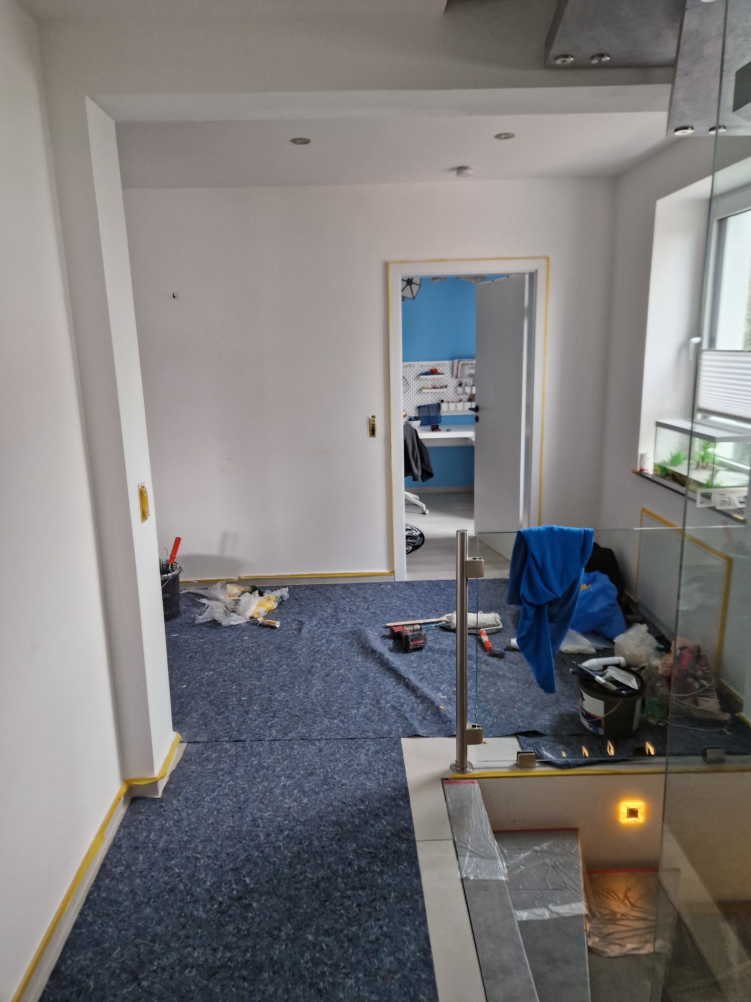 Wnętrze w trakcie prac wykończeniowych: zabezpieczona podłoga, ściany przygotowane do malowania, widoczne narzędzia malarskie i drabina w tle, pomieszczenie z niebieską ścianą widoczne przez...