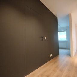 Fix&Finish Robert Tomala - Ciemnoszara ściana z ukrytymi drzwiami i minimalistyczną klamką, widoczny fragment podłogi z paneli oraz przejście do jasnego pomieszczenia z oknem i grzejnikiem.