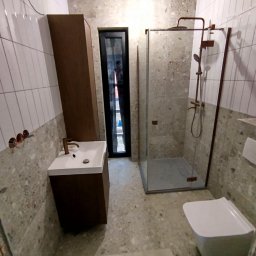 Fix&Finish Robert Tomala - Nowoczesna łazienka z kabiną prysznicową, umywalką z szafką w kolorze drewna i toaletą, wykończona płytkami imitującymi kamień i białymi płytkami na ścianach.