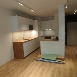Fix&Finish Robert Tomala - Nowoczesna, jasna kuchnia z drewnianym blatem i białymi szafkami. Na podłodze panele, a w tle widoczne wejście do innego pomieszczenia. Częściowo rozpakowane panele na podłodze.