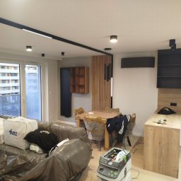 Fix&Finish Robert Tomala - Jasne wnętrze po remoncie: nowoczesne oświetlenie, drewniane akcenty, meble zabezpieczone folią, sprzęt budowlany. Widok na okno i zabudowę kuchenną.