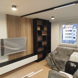 Fix&Finish Robert Tomala - Nowoczesny salon w Poznaniu z drewnianą ścianą, czarnym regałem i listwą LED. Telewizor zabezpieczony folią, podłoga częściowo zakryta. Minimalistyczny design.