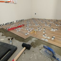Fix&Finish Robert Tomala - Układanie drewnopodobnych płytek podłogowych z użyciem systemu poziomowania. Widoczne narzędzia: poziomica, młotek gumowy, kielnie. Jasne wnętrze w trakcie prac.