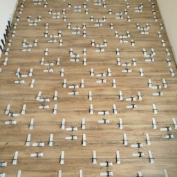 Fix&Finish Robert Tomala - Ułożone panele podłogowe z systemem poziomowania, widok z góry, jasne drewno, szare fugi, białe klipsy, czarne kliny przy ścianie, fragment buta w rogu.