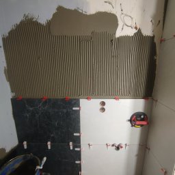 Fix&Finish Robert Tomala - Proces układania płytek w łazience: widoczna nałożona zaprawa, częściowo ułożone czarne i białe płytki, przyssawka do płytek i system poziomowania płytek.