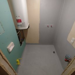 Fix&Finish Robert Tomala - Wnętrze łazienki w trakcie prac: ściany częściowo pomalowane na szaro, widoczny bojler, płyta gipsowa i instalacje. Podłoga w szarym kolorze, widoczne elementy instalacji.