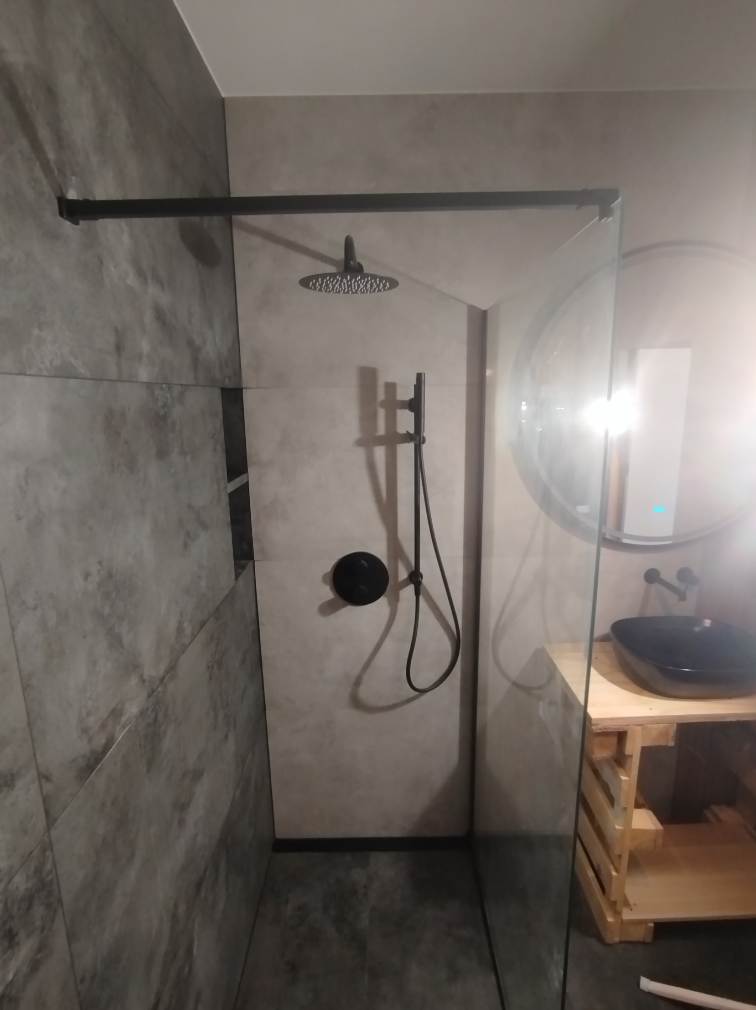 Nowoczesna kabina prysznicowa walk-in z czarną armaturą, deszczownicą i szklaną ścianką, umywalka nablatowa na drewnianej konstrukcji przypominającej paletę, szare płytki imitujące beton.