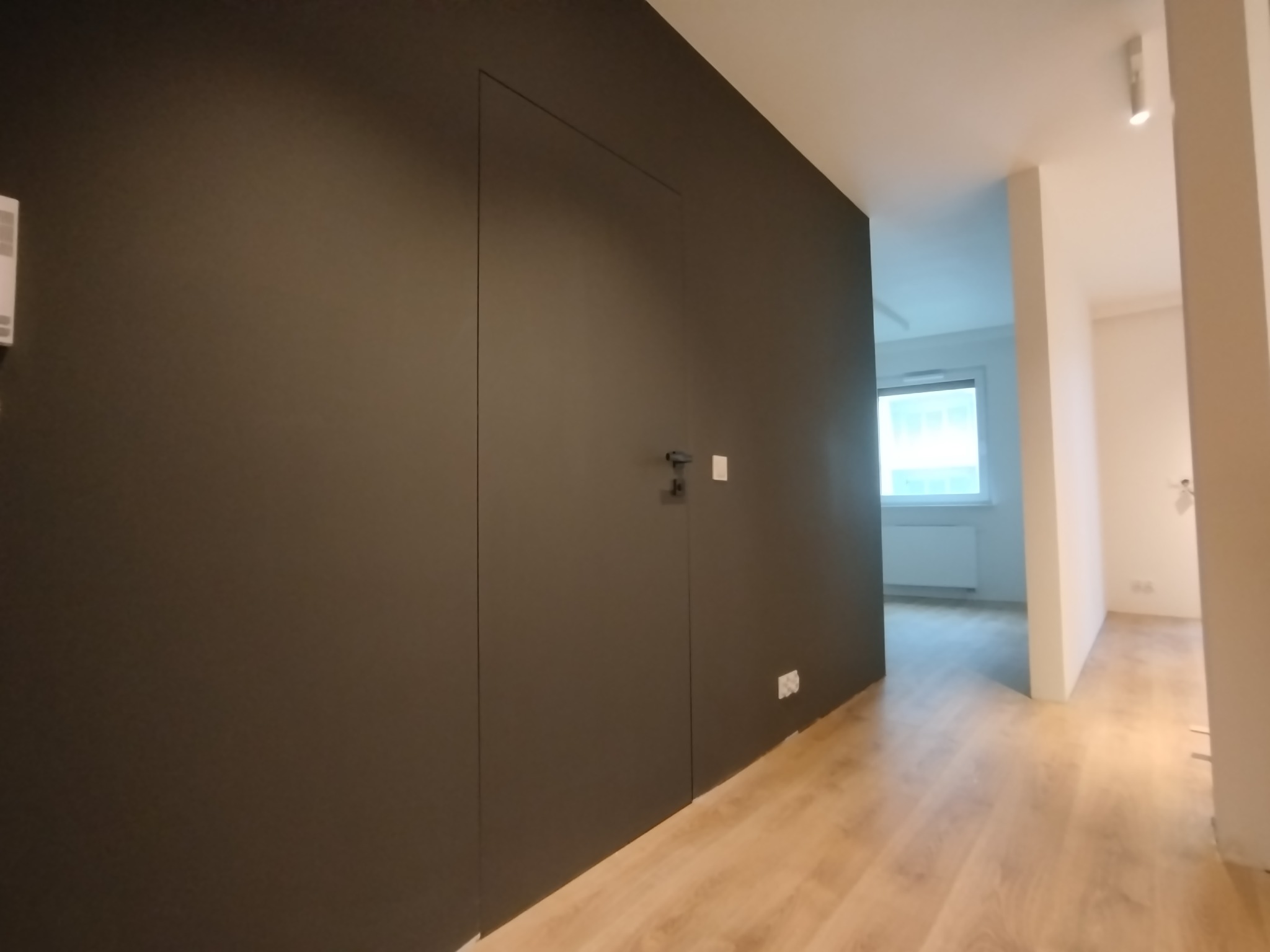 Ciemnoszara ściana z ukrytymi drzwiami i minimalistyczną klamką, widoczny fragment podłogi z paneli oraz przejście do jasnego pomieszczenia z oknem i grzejnikiem.