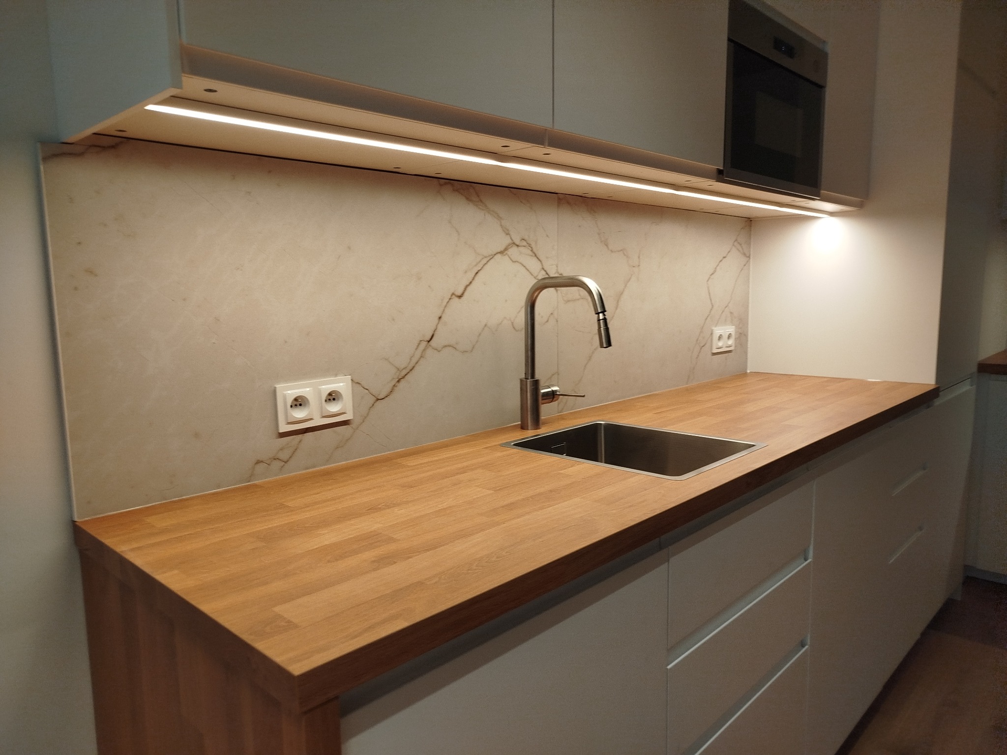 Nowoczesna kuchnia z drewnianym blatem, marmurową ścianą nad blatem, oświetleniem LED pod szafkami i minimalistycznym zlewem ze stali nierdzewnej.