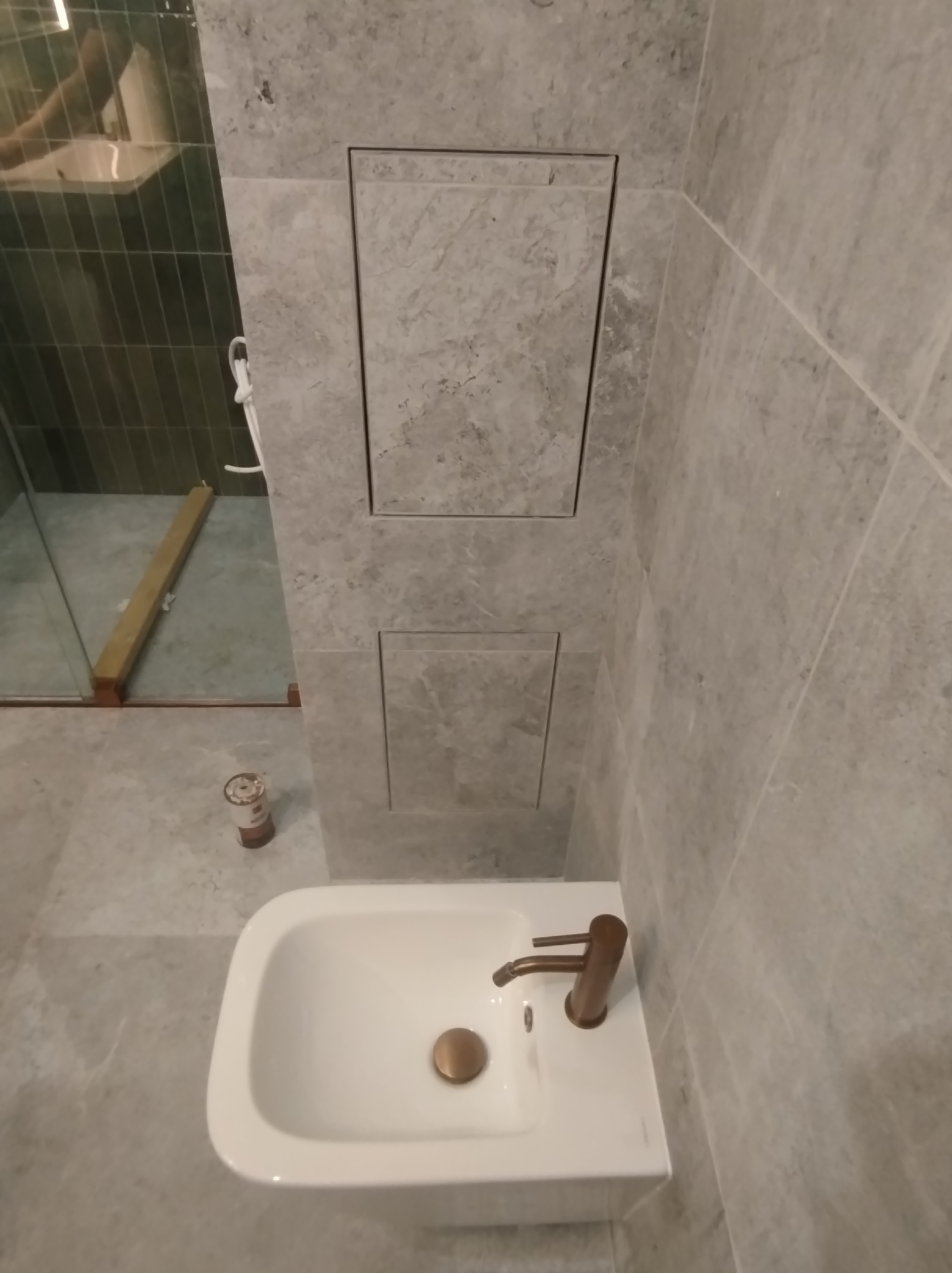 Nowoczesna biała umywalka z miedzianą armaturą na tle szarych płytek imitujących marmur, z ukrytymi drzwiczkami rewizyjnymi w ścianie i widoczną kabiną prysznicową w tle.