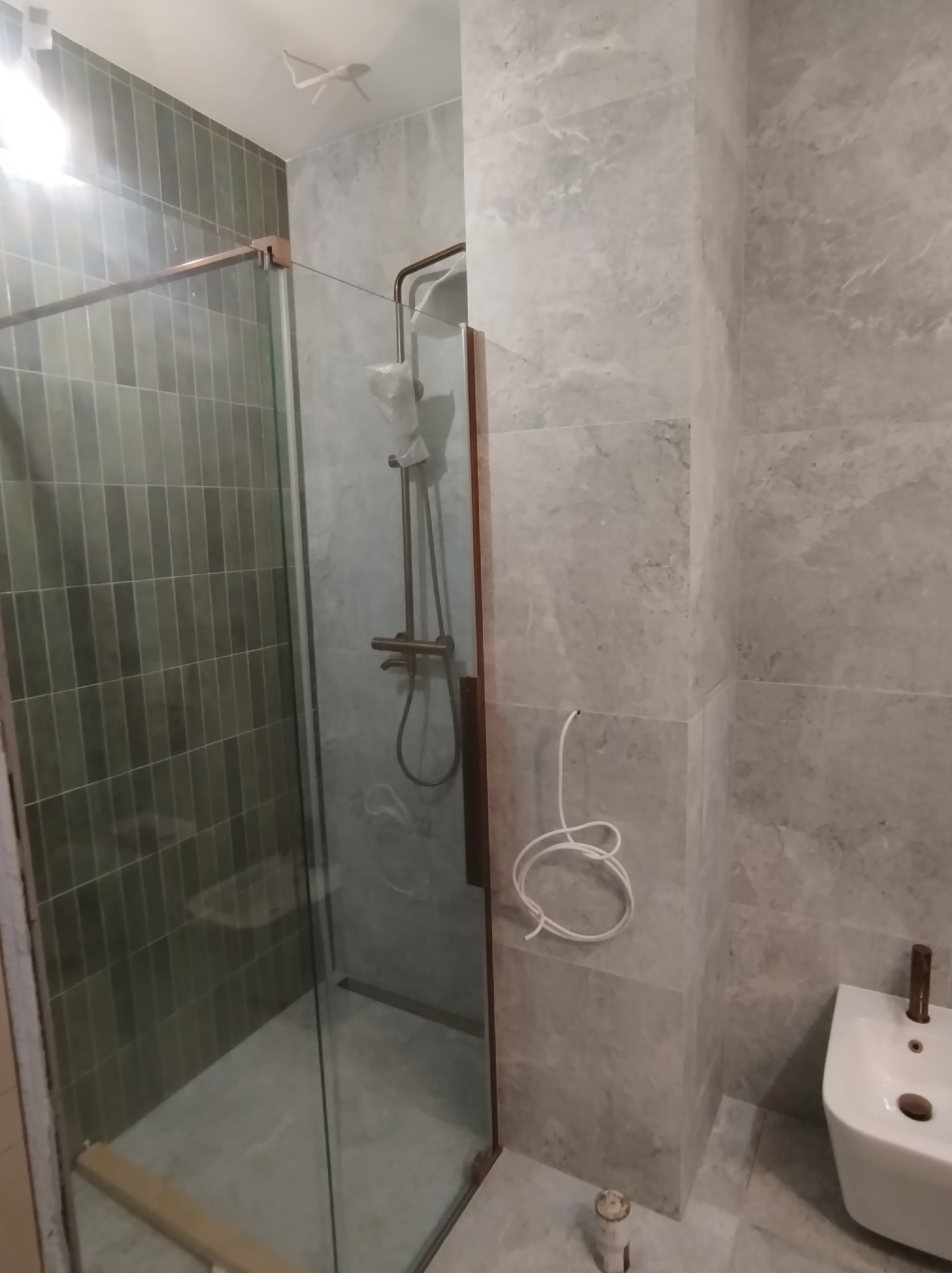 Nowoczesna łazienka w trakcie wykańczania: kabina prysznicowa ze złotymi elementami, białe bidet, szare płytki imitujące marmur, zwisający kabel elektryczny.