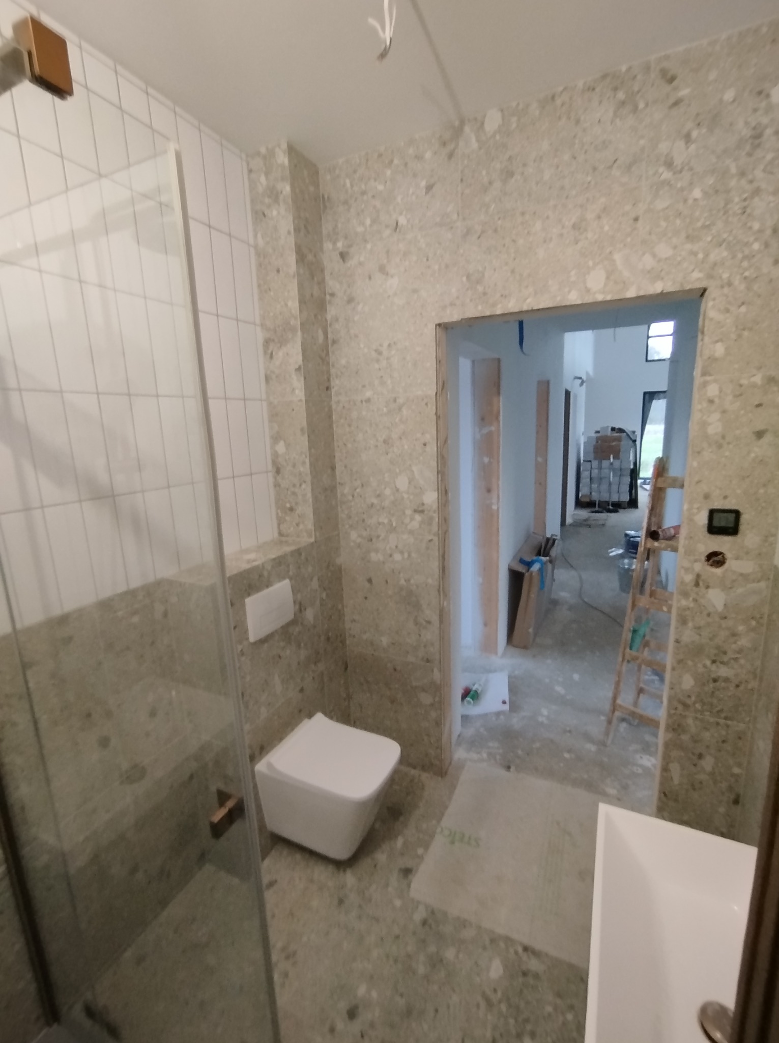 Wnętrze łazienki w trakcie remontu, widoczne płytki imitujące lastryko, kabina prysznicowa ze szklanymi drzwiami, podwieszana toaleta, oraz widok na dalszą część domu z drabiną i materiałami...