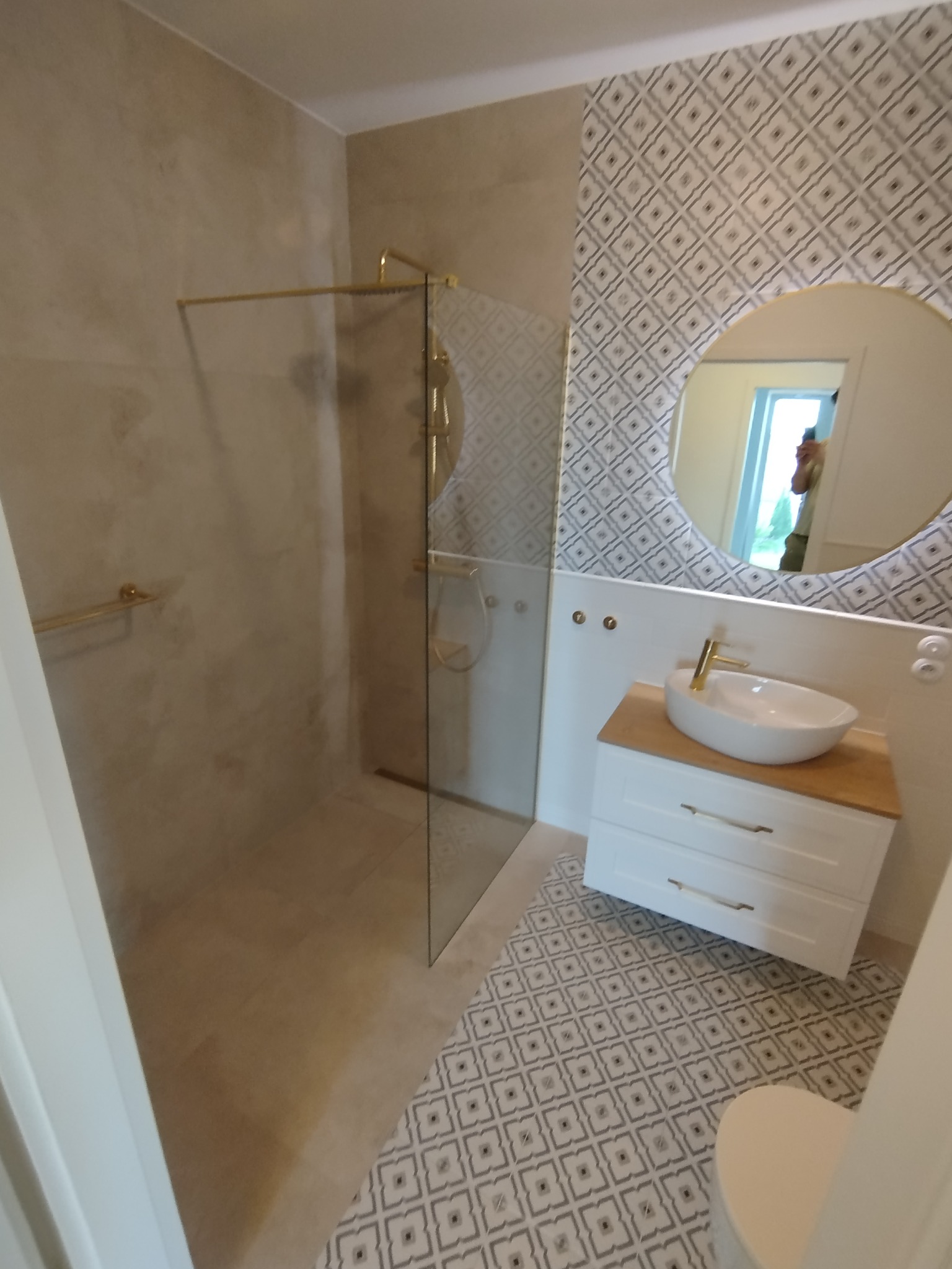 Nowoczesna łazienka z kabiną prysznicową ze złotymi elementami, umywalką na szafce z drewnianym blatem i lustrem w okrągłej ramie.