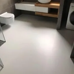 Jasnoszara, jednolita podłoga w nowoczesnej łazience z wiszącą miską WC, umywalką nablatową na drewnianym blacie i pralką. Powierzchnia podłogi gładka, bez spoin.