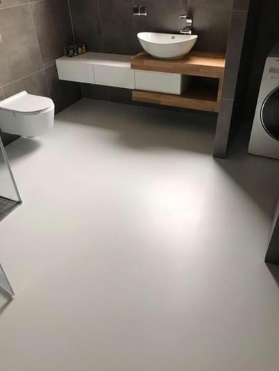 Jasnoszara, jednolita podłoga w nowoczesnej łazience z wiszącą miską WC, umywalką nablatową na drewnianym blacie i pralką. Powierzchnia podłogi gładka, bez spoin.