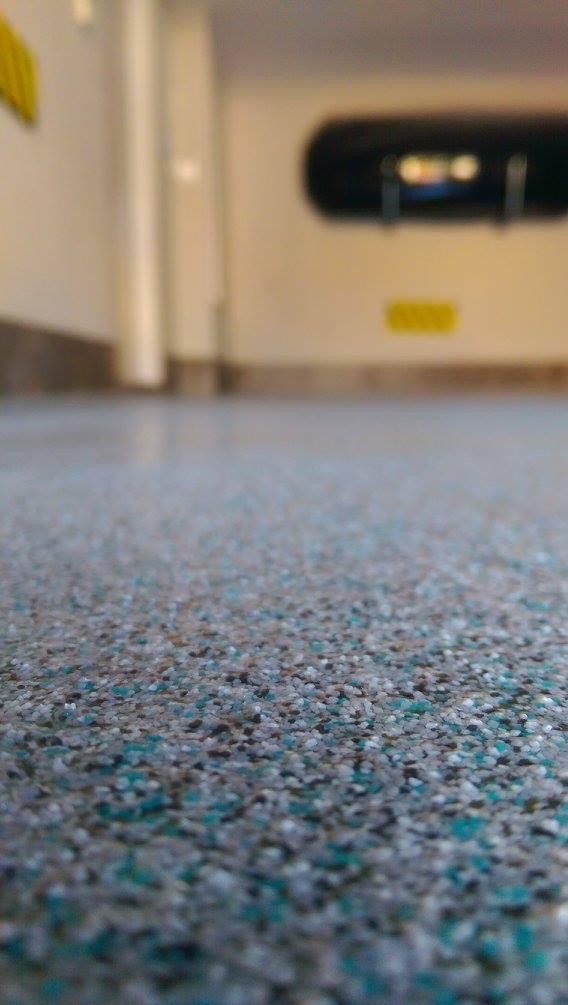 Szara posadzka z drobnymi, kolorowymi elementami, widok z niskiej perspektywy, z rozmytym tłem ściany i elementami wyposażenia.