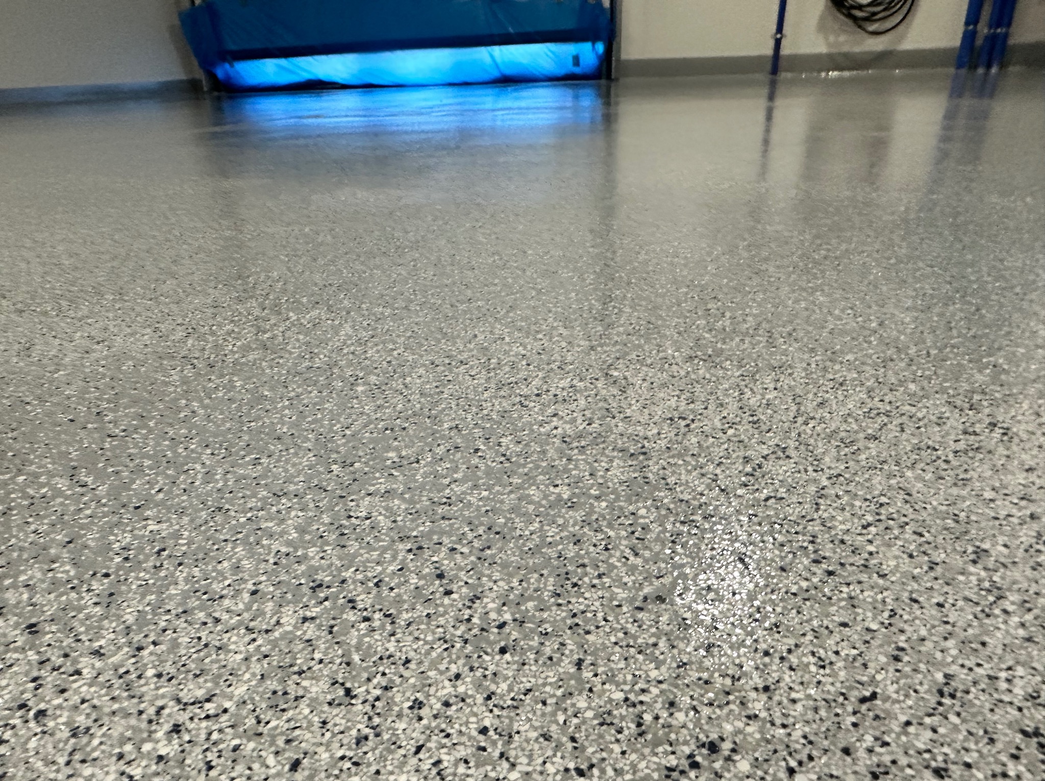 Jasnoszara posadzka żywiczna z drobnymi czarnymi i białymi wtrąceniami, gładka powierzchnia odbija światło, w tle widoczna brama garażowa z niebieskim uszczelnieniem.